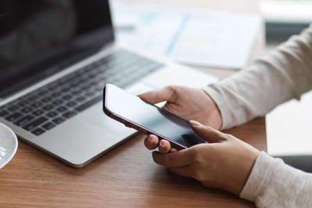 Close-up side view of female hands using smartphone with laptop, papers work on the wooden deskの写真素材