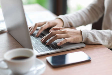 Close up of female fingers typing on laptop keyboard with smartphone and coffee on the wooden deskの写真素材