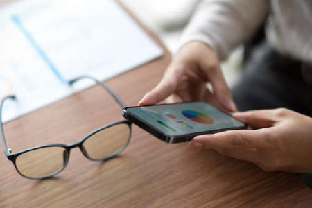 Close up of female looking on financial chart on her smartphone with eye-glasses and financial report on wooden tableの写真素材