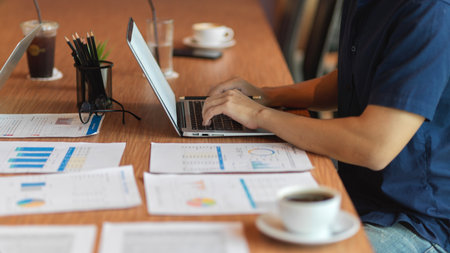 Businessman working on laptop with financial report, office supplies, coffee cup on the wooden deskの写真素材