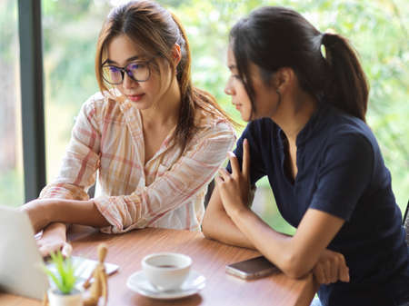 Portrait of 2 businesswomen discussing and looking on laptop with mobile phone and coffee cup on wooden deskの写真素材
