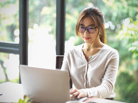 Portrait of beautiful asian businesswoman with eyeglasses working on laptop at office with blurred natural in the backgroundの写真素材