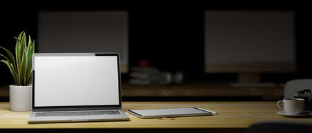 Work late concept, Portable laptop blank screen with tablet mock up on wood table in low light office interior in the background, 3d rendering, 3d illustrationの写真素材