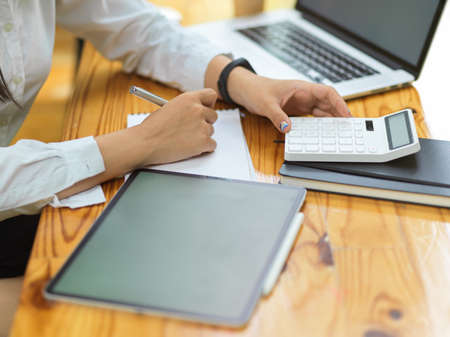 Close up female hands working with laptop, tablet and calculator on wooden table, Office concept, Calculating company expensesの写真素材