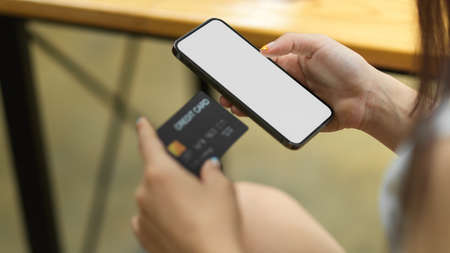 Close up female hand hold credit card and smartphone with blank empty screen and blurred background,...
