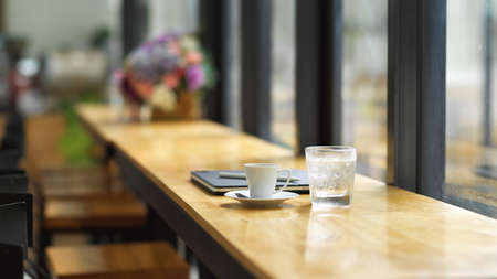 Cafe working space, with a glass of ice water, coffee cup and portable laptop on wooden table with cafe interior in the backgroundの写真素材