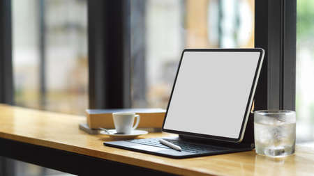 Freelancer lifestyle, Blank screen tablet with keyboard and copy space on wooden table with coffee cup, a glass of ice water and book, work via internet during coffee breakの写真素材