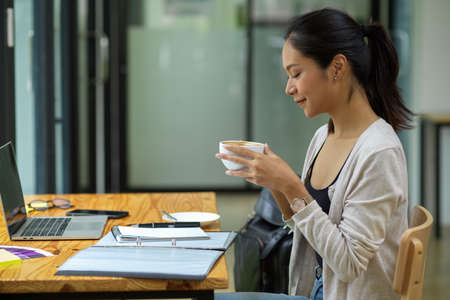 Female manager drinking coffee, feeling happy during reading financial annual net profit with opened laptop, report and stuffの写真素材