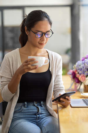 Attractive asian female with eye-glasses and casual clothes drinking hot coffee while looking at her smartphone, freelancer weekend's work at coffee shopの写真素材