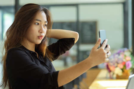 Attractive female taking pic of herself by her smartphone, Beauty asian brunette girl taking selfie, blurred backgroundの写真素材