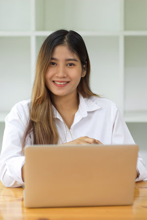 Portrait of beautiful female in white long sleeve shirt looks sitting in front of computer laptop, business female portraitの写真素材