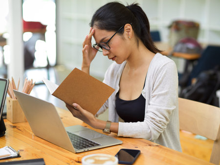 Stressed casual female with glasses worker looking on her schedule book with laptop, stationery and phone on desk at officeの写真素材