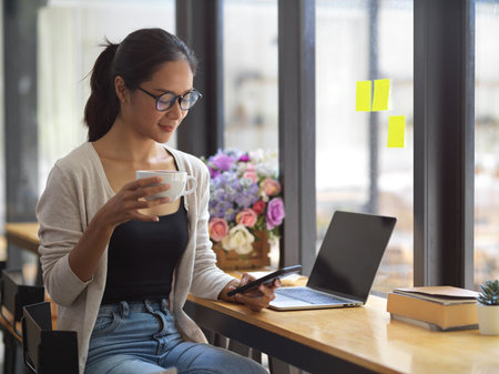 Attractive female freelancer with eye-glasses drinking coffee and scrolling on smartphone with opened laptop at cafeの写真素材
