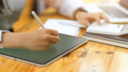 Cropped shot of female hands using tablet and stylus pen while calculating on wooden table