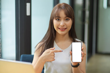 Portrait of happy female hold smart phone with blank empty screen with office interior in the backgroundの写真素材
