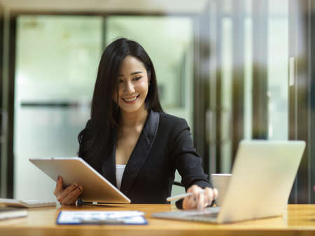 Attractive businesswoman in black suit, using laptop and holding smart tablet to check business...