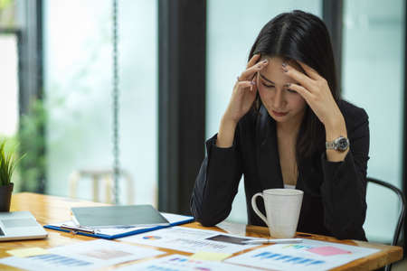 Attractive businesswoman getting stressed out from work with financial reports, tablet, laptop, coffee cup on wood desk at officeの写真素材