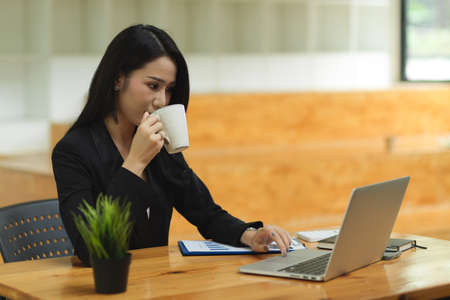 Confident businesswoman drinking  coffee during working on laptop, smart businesswoman, female in black suit, at officeの写真素材
