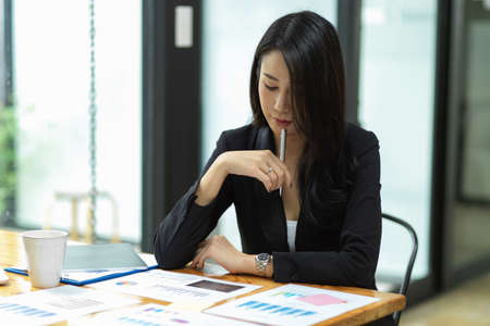 Thoughtful businesswoman thinking idea to solve business problem with financial report, tablet, glass of water on desk at office, making decisionの写真素材
