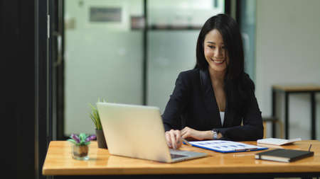 Beautiful businesswoman looking, checking, working on laptop with smile face, office supplies on wooden table, office work space, girl in black suitの写真素材