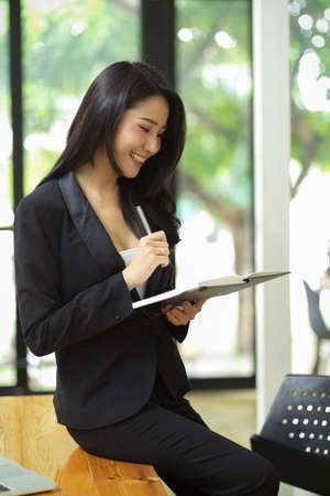 Elegant businesswoman in black suit stands leaning on desk with holding planner book at office, smiling millennial office managerの写真素材