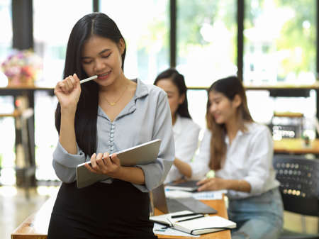 Attractive asian company employee stands in front of worktable and think about business work with digital tablet, thoughtful woman, business partners in backgroundの写真素材