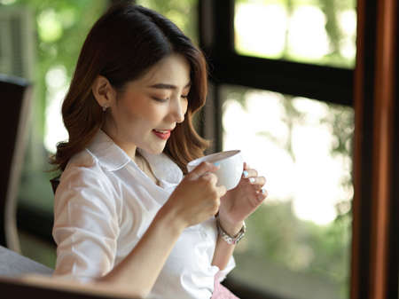 Young asian working woman enjoy her hot coffee at coffee shop, sipping morning coffee, blur bokeh backgroundの写真素材