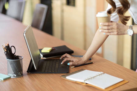 Close up, side view, business female lean on office desk, typing on tablet keyboard while holding take away coffee at officeの写真素材