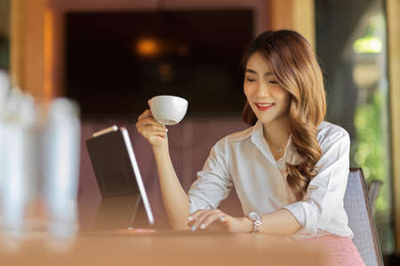 Happy beautiful businesswoman working, answering message on tablet and drinking morning coffee at office, relaxing with good hot coffeeの写真素材