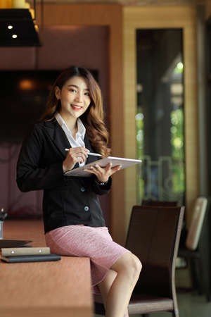 Charming business female sitting on her office desk, holding using tablet computer and stylus penの写真素材