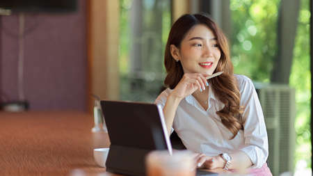 Attractive businesswoman looking away and thinking about her business project idea, tablet on wood table at officeの写真素材