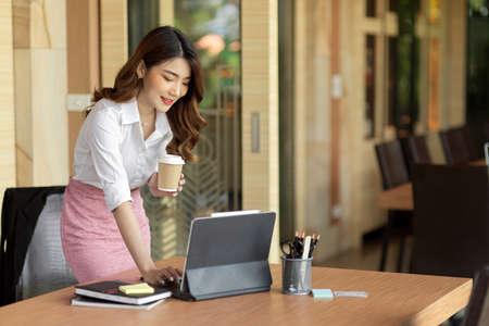 Professional executive businesswoman standing, holding take away coffee and working on digital tablet at her office deskの写真素材