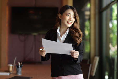 Businesswoman looking away through the window, smiling, thinking about business plan, holding business papers at her officeの写真素材