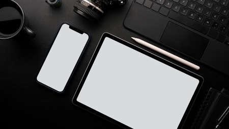 Photographer work table concept, blank screen tablet and smartphone mockup with film camera, laptop, coffee on black backgroundの写真素材
