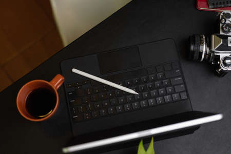 Top view on modern workplace of photographer, portable tablet with keyboard, camera, coffee mug, stylus pen on black backgroundの写真素材