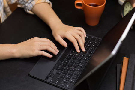 Female college student doing homework on smart tablet, online class, typing on tablet keyboard, modern black workspace, close up handsの写真素材