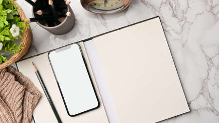 Workspace in autumn composition, smartphone mockup, notebook blank space for your text on marble background, vintage alarm clock, pencils, sweater, plant, flat layの写真素材