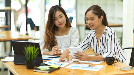 Colleague in office concept, two casual asian businesswomen discussing work in office, laptop, financial papers, calculator, office backgroundの写真素材
