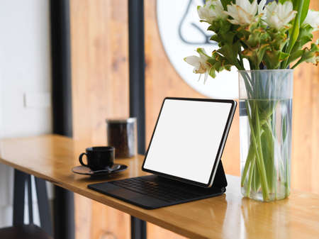 Portable workspace with portable tablet blank screen mockup with magic keyboard, coffee cup, flowers in glass vase on wood tableの写真素材