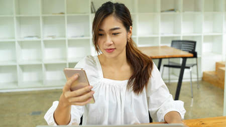 Pretty woman using smartphone texting to her friends, scrolling on social media, co-working space in the backgroundの写真素材