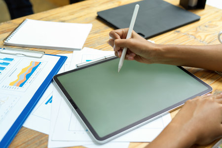 Closeup hand, holding stylus pen, writing on smart tablet, tablet blank screen mockup, financial paperwork on tableの写真素材