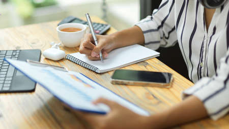 Businesswoman taking notes, planning business model from financial paperwork, laptop, tea cup, calculator on wood table, cropped shotの写真素材
