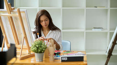 Young gorgeous female artist looking at oil colour for her art work, sit in front of canvas easel at studioの写真素材
