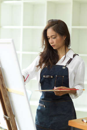 Attractive asian female artist working in her studio, passion painting, masterpiece, holding art palette, paint brush in handの写真素材