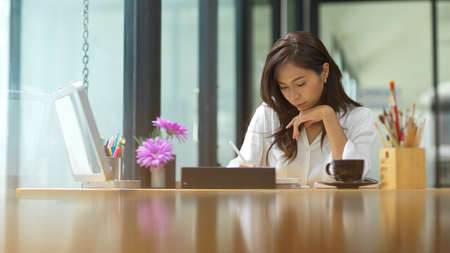 Professional female graphic art designer working on smart tablet in her office, painting tools and coffee mug on deskの写真素材