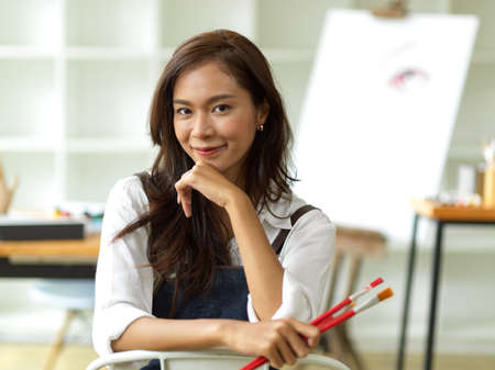Beautiful asian female artist image, holding painting brushes, sitting, hand on chin, blurred art work studio in the backgroundの写真素材