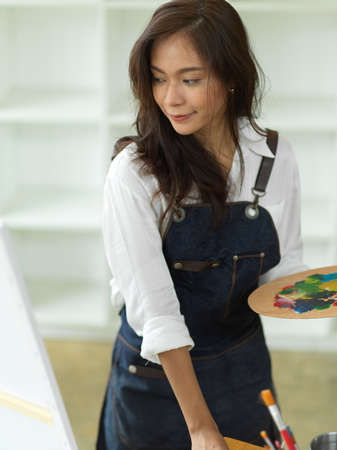 Portrait of female painting on canvas easel, holding colour palette, painting in art workshopの写真素材