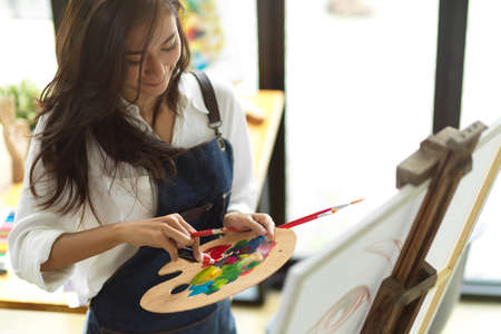 Young female student doing some artwork with acrylic colour, painting supplies, holding palette, creative hobbyの写真素材