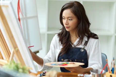 Professional female artist painting on canvas easel, holding colour palette at art studio, creative spaceの写真素材