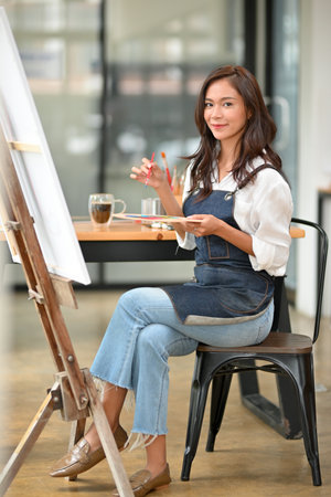 Portrait of attractive young female holding palette colour, large canvas easel in her art studioの写真素材